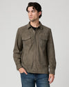 A man wearing a dark brown suede long-sleeve button-up shirt with two chest pockets, paired with blue jeans, photographed against a neutral light gray background.