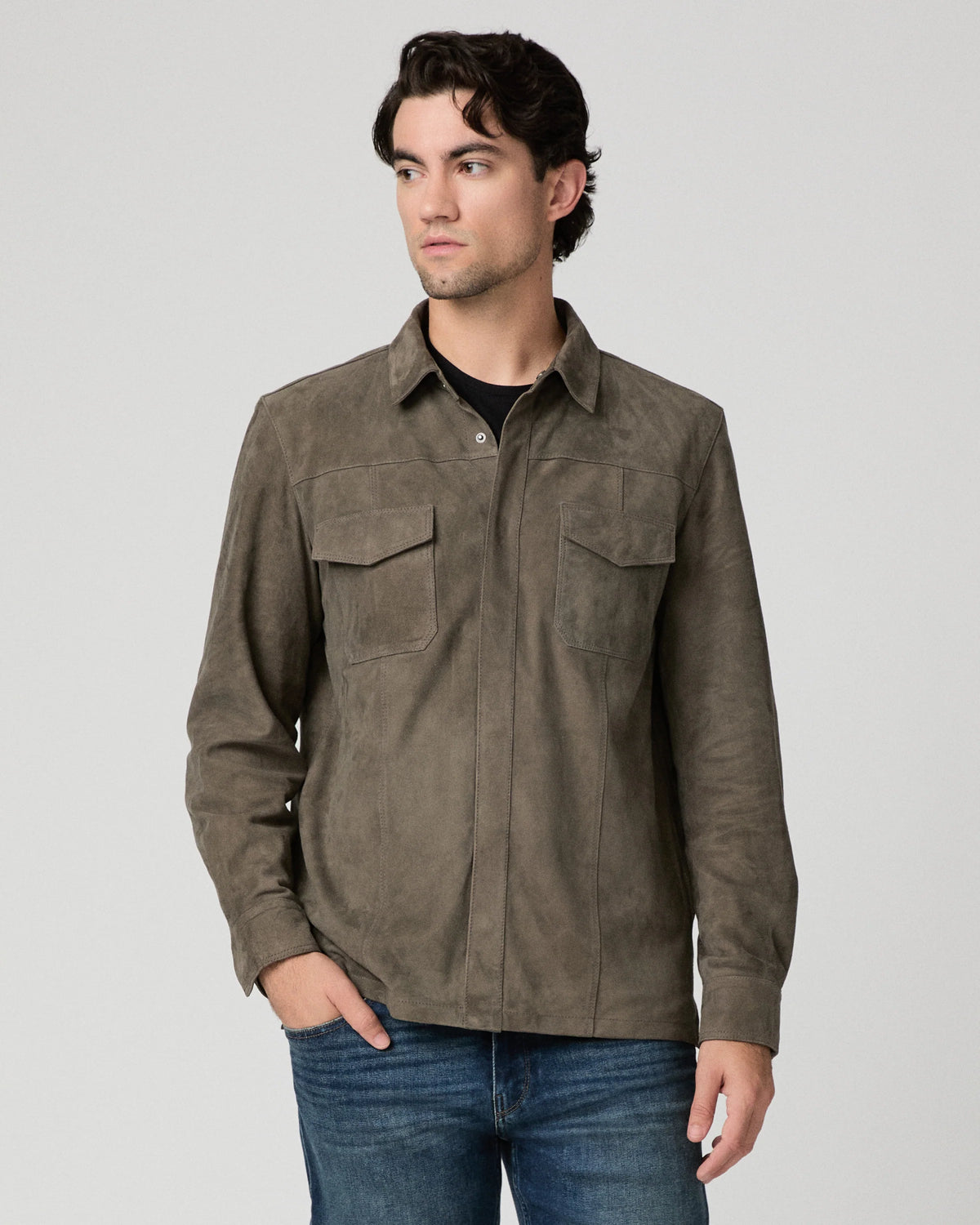 A man wearing a dark brown suede long-sleeve button-up shirt with two chest pockets, paired with blue jeans, photographed against a neutral light gray background.