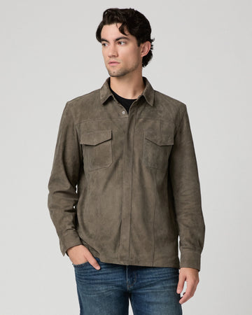 A man wearing a dark brown suede long-sleeve button-up shirt with two chest pockets, paired with blue jeans, photographed against a neutral light gray background.