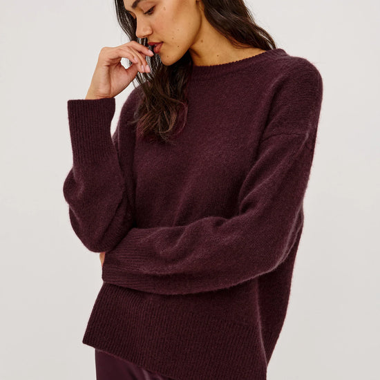 A woman wearing a fuzzy, oversized crewneck sweater in deep plum purple, paired with a matching satin skirt, against a plain light gray studio background.