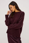 A woman wearing a fuzzy, oversized crewneck sweater in deep plum purple, paired with a matching satin skirt, against a plain light gray studio background.