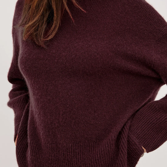 A close-up of a woman wearing a chunky knit crewneck sweater in a deep plum purple color, paired with a matching dark skirt against a plain light background.