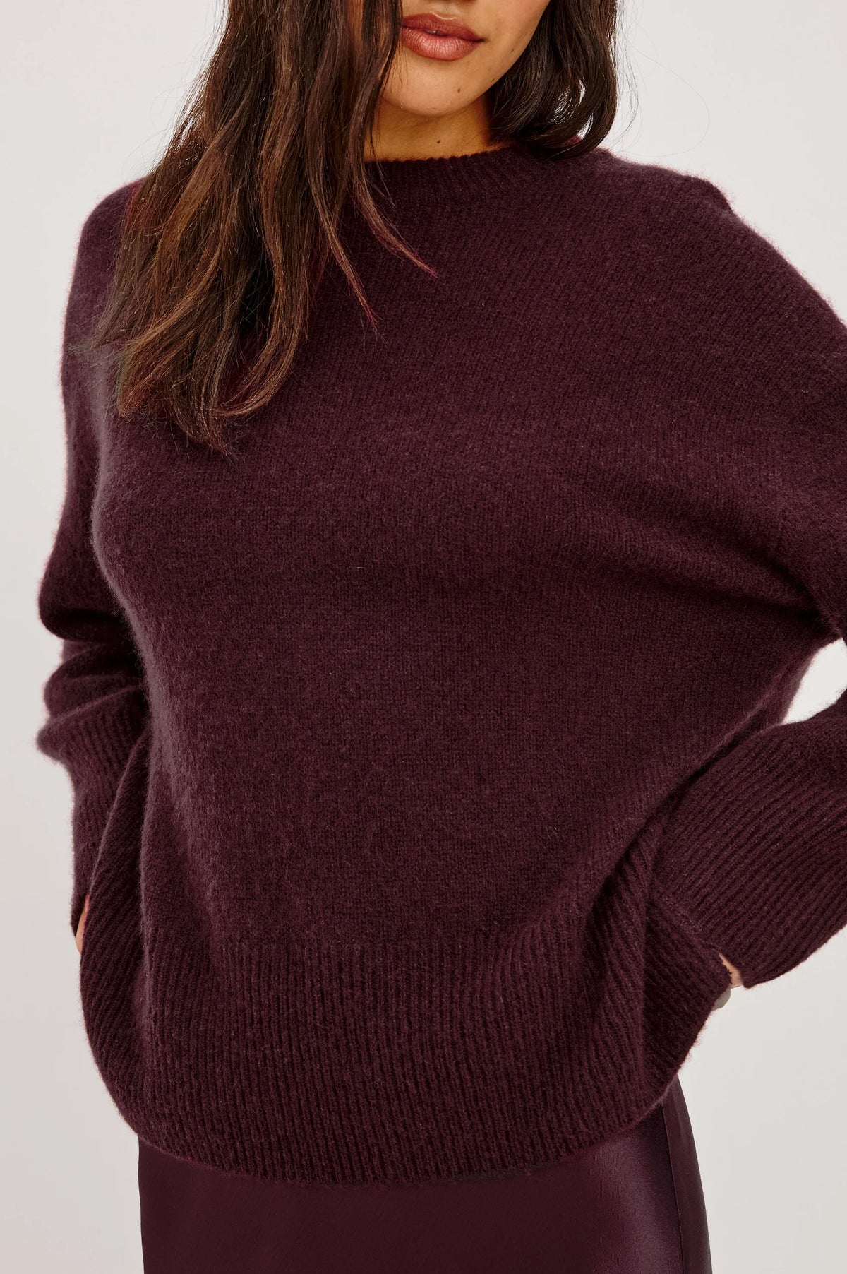 A close-up of a woman wearing a chunky knit crewneck sweater in a deep plum purple color, paired with a matching dark skirt against a plain light background.