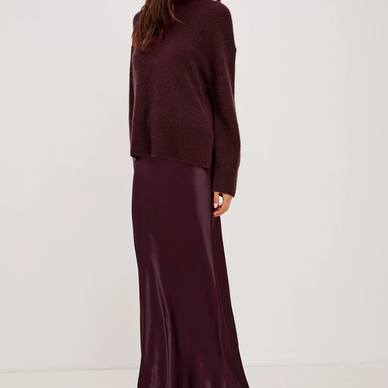 A woman modeling a matching two-piece outfit consisting of a dark plum knit sweater and a long satin maxi skirt, set against a plain light gray studio background.