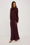 A woman modeling a matching two-piece outfit consisting of a dark plum knit sweater and a long satin maxi skirt, set against a plain light gray studio background.