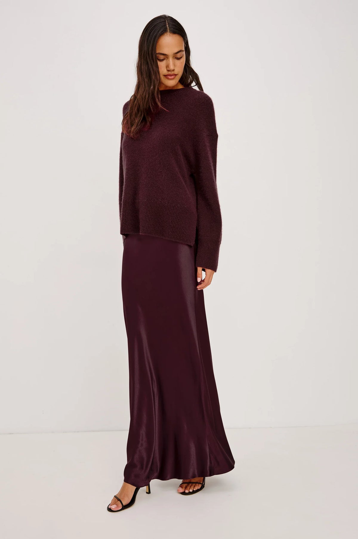 A woman modeling a matching two-piece outfit consisting of a dark plum knit sweater and a long satin maxi skirt, set against a plain light gray studio background.