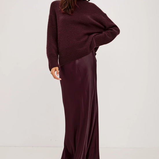 A woman modeling a deep burgundy two-piece outfit consisting of a chunky knit long-sleeve sweater and a matching satin maxi skirt, paired with black strappy sandals against a plain white studio background.