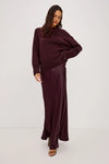 A woman modeling a deep burgundy two-piece outfit consisting of a chunky knit long-sleeve sweater and a matching satin maxi skirt, paired with black strappy sandals against a plain white studio background.