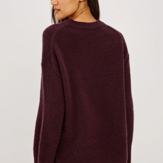 A woman shown from the back wearing a dark plum-colored knit sweater and matching skirt against a plain light gray studio background.