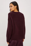 A woman shown from the back wearing a dark plum-colored knit sweater and matching skirt against a plain light gray studio background.