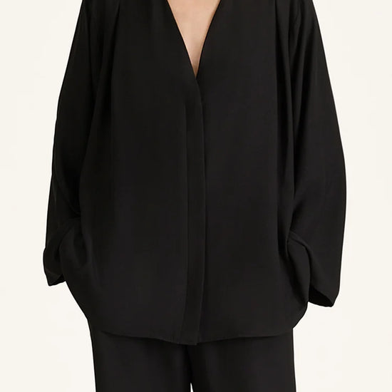 A model wearing a black draped long-sleeve wrap jacket with a deep V-neckline, paired with matching black trousers, against a plain off-white background.