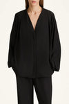 A model wearing a black draped long-sleeve wrap jacket with a deep V-neckline, paired with matching black trousers, against a plain off-white background.