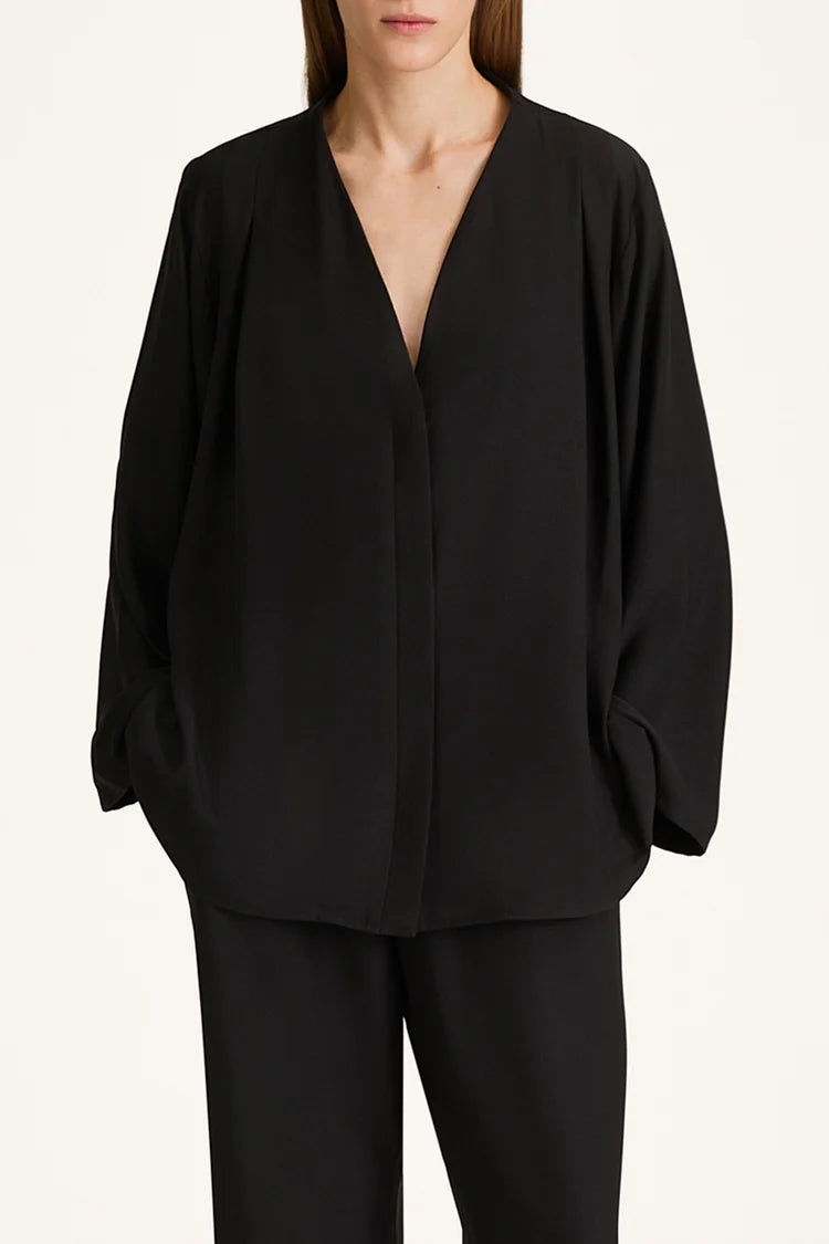 A model wearing a black draped long-sleeve wrap jacket with a deep V-neckline, paired with matching black trousers, against a plain off-white background.