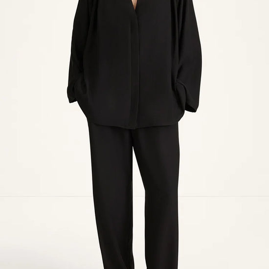 A model wearing a black draped two-piece set consisting of a long-sleeve V-neck top and matching wide-leg trousers, photographed against a clean white studio background.