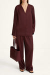 A model wearing a matching burgundy two-piece set consisting of a long-sleeve V-neck top and wide-leg trousers, holding a matching burgundy tote bag against a plain white studio background.