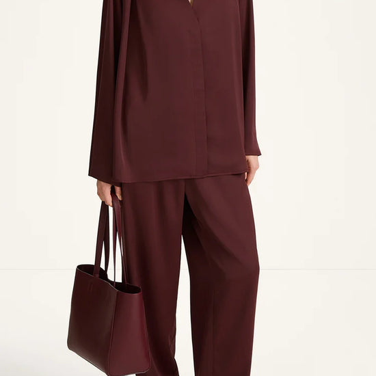 A model wearing a matching burgundy two-piece set consisting of a long-sleeve wrap-style top and wide-leg trousers, holding a matching burgundy tote bag against a plain white studio background.