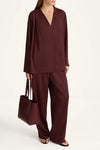 A model wearing a matching burgundy two-piece set consisting of a long-sleeve wrap-style top and wide-leg trousers, holding a matching burgundy tote bag against a plain white studio background.