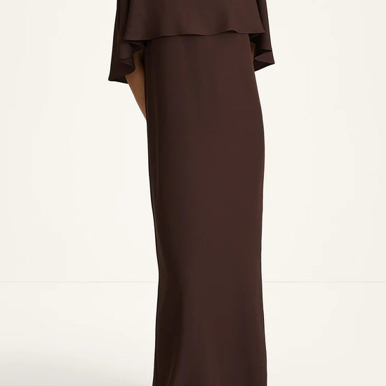 A woman modeling a floor-length dark brown dress with a layered ruffle neckline and short flutter sleeves, photographed against a plain off-white studio background.