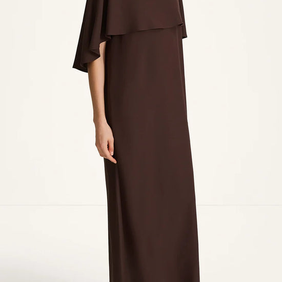 A woman modeling a dark brown, floor-length maxi dress with draped cap sleeves and a layered ruffle detail across the chest, paired with black flat sandals against a plain white studio background.