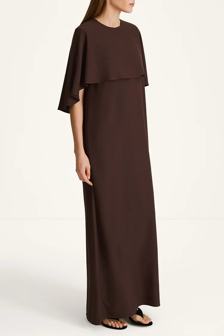 A woman modeling a dark brown, floor-length maxi dress with draped cap sleeves and a layered ruffle detail across the chest, paired with black flat sandals against a plain white studio background.
