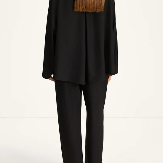 A model shown from the back wearing a black draped long-sleeve blazer and matching wide-leg trousers against a clean white studio background.