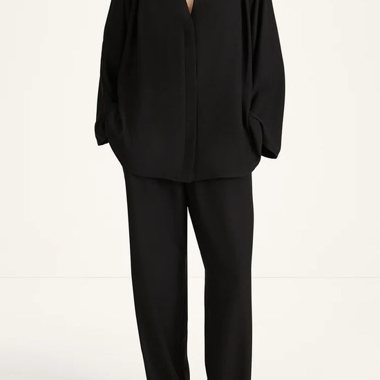 A model wearing a black draped two-piece set consisting of a long-sleeve V-neck top and matching wide-leg trousers, photographed against a clean white studio background.