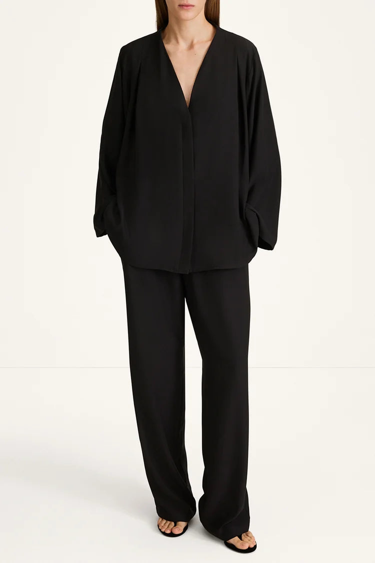 A model wearing a black draped two-piece set consisting of a long-sleeve V-neck top and matching wide-leg trousers, photographed against a clean white studio background.