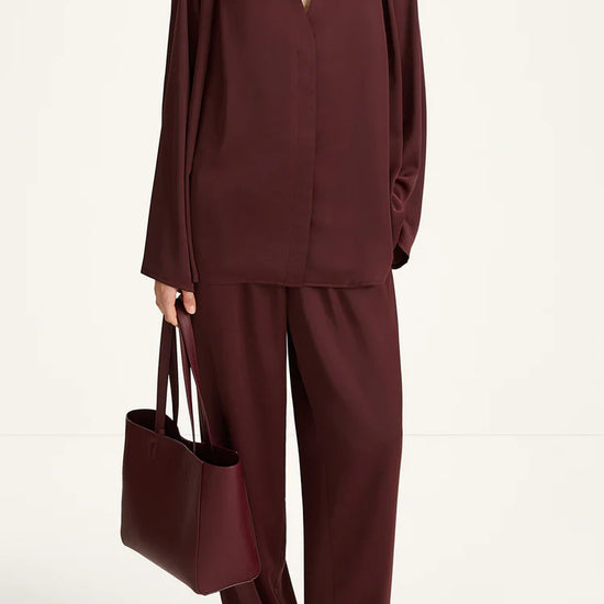 A model wearing a matching burgundy two-piece set consisting of a long-sleeve V-neck top and wide-leg trousers, holding a matching burgundy tote bag against a plain white studio background.