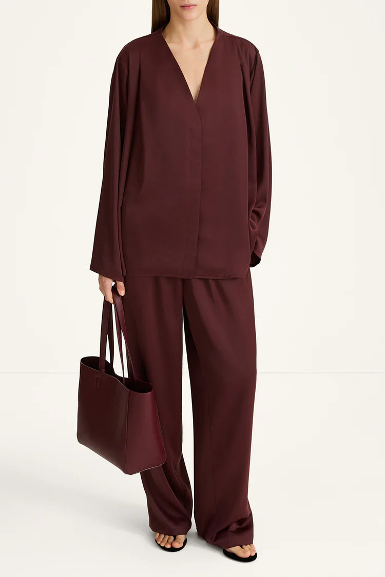 A model wearing a matching burgundy two-piece set consisting of a long-sleeve V-neck top and wide-leg trousers, holding a matching burgundy tote bag against a plain white studio background.