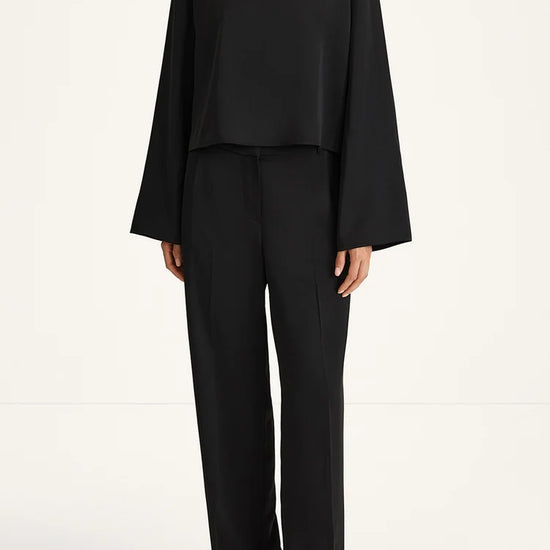 A model wearing a black two-piece outfit consisting of a long-sleeve cropped top and matching wide-leg trousers, paired with black sandals, against a plain white studio background.