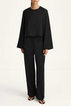 A model wearing a black two-piece outfit consisting of a long-sleeve cropped top and matching wide-leg trousers, paired with black sandals, against a plain white studio background.