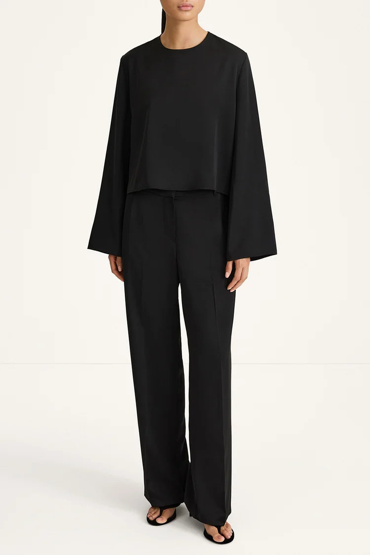 A model wearing a black two-piece outfit consisting of a long-sleeve cropped top and matching wide-leg trousers, paired with black sandals, against a plain white studio background.