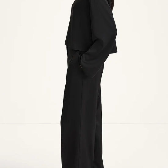 A model shown in profile wearing a black two-piece outfit consisting of a draped long-sleeve top and matching wide-leg trousers, paired with black high-heeled sandals against a plain white studio background.