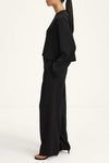 A model shown in profile wearing a black two-piece outfit consisting of a draped long-sleeve top and matching wide-leg trousers, paired with black high-heeled sandals against a plain white studio background.