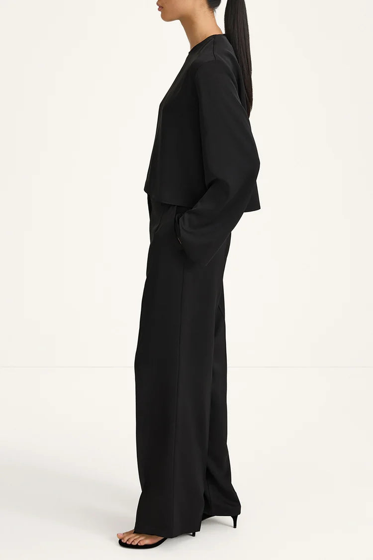 A model shown in profile wearing a black two-piece outfit consisting of a draped long-sleeve top and matching wide-leg trousers, paired with black high-heeled sandals against a plain white studio background.