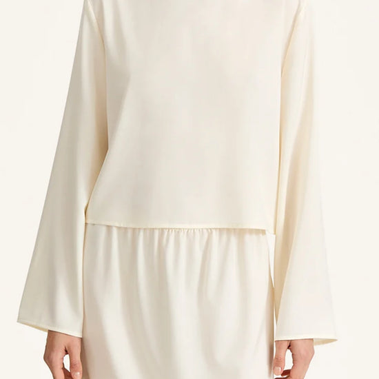 A model wearing a cream-colored long-sleeve top with a matching skirt, photographed against a clean, light off-white background.
