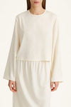 A model wearing a cream-colored long-sleeve top with a matching skirt, photographed against a clean, light off-white background.