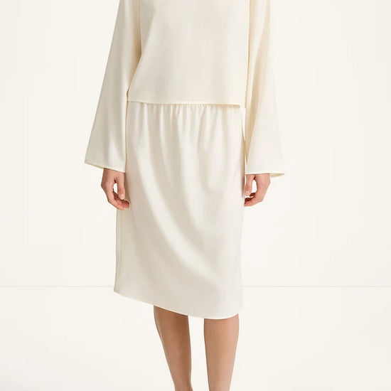 A model wearing a cream-colored two-piece set consisting of a long-sleeve cropped top and a matching knee-length skirt, paired with black flip-flop sandals against a plain white studio background.