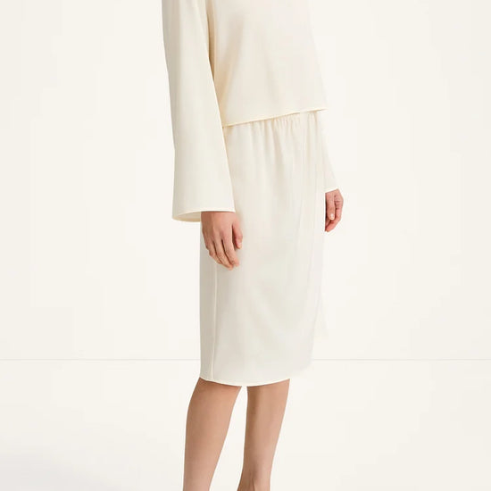 A model wearing a cream-colored two-piece set consisting of a long-sleeve cropped top and a matching midi skirt, paired with black heeled sandals against a plain white studio background.
