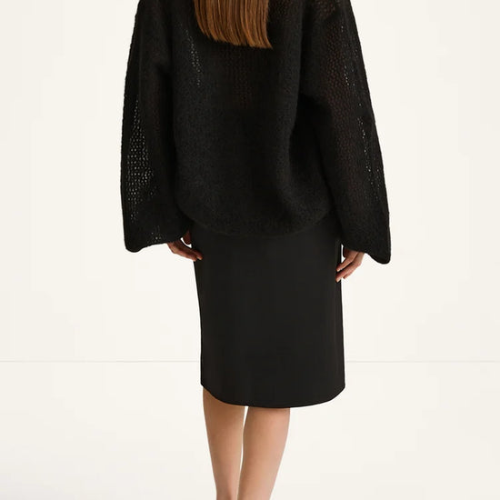 A model shown from the back wearing a black open-knit cropped sweater and a black midi skirt, paired with black mule sandals, against a plain white studio background.