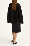 A model shown from the back wearing a black open-knit cropped sweater and a black midi skirt, paired with black mule sandals, against a plain white studio background.