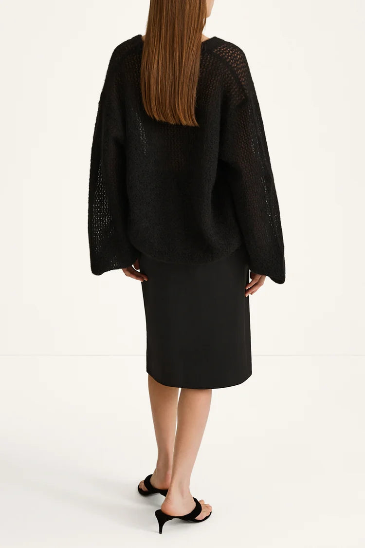 A model shown from the back wearing a black open-knit cropped sweater and a black midi skirt, paired with black mule sandals, against a plain white studio background.