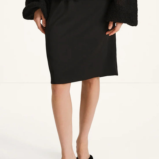 A model wearing a black knee-length skirt, a black textured cardigan, and black strappy heeled sandals, shown from the chest down against a plain white studio background.