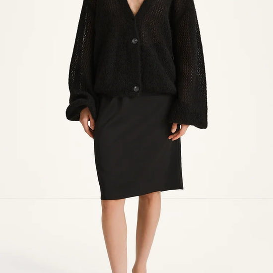 A model wearing a black open-knit cardigan with long balloon sleeves and a black pencil skirt, paired with black strappy sandals, against a plain white studio background.
