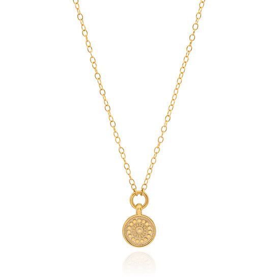 A delicate gold chain necklace with a small round pendant featuring an intricate floral or sunburst pattern, displayed on a clean white background with a subtle reflection.