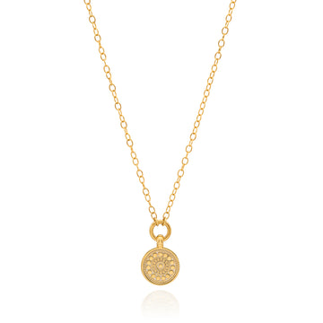 A delicate gold chain necklace with a small round pendant featuring an intricate floral or sunburst pattern, displayed on a clean white background with a subtle reflection.