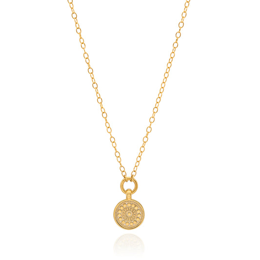 A delicate gold chain necklace with a small round pendant featuring an intricate floral or sunburst pattern, displayed on a clean white background with a subtle reflection.