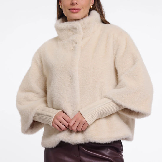 A woman wearing a cream-colored, fluffy faux-fur cropped jacket with a high mock neck and ribbed knit sleeves, paired with dark brown leather pants, against a plain white studio background.