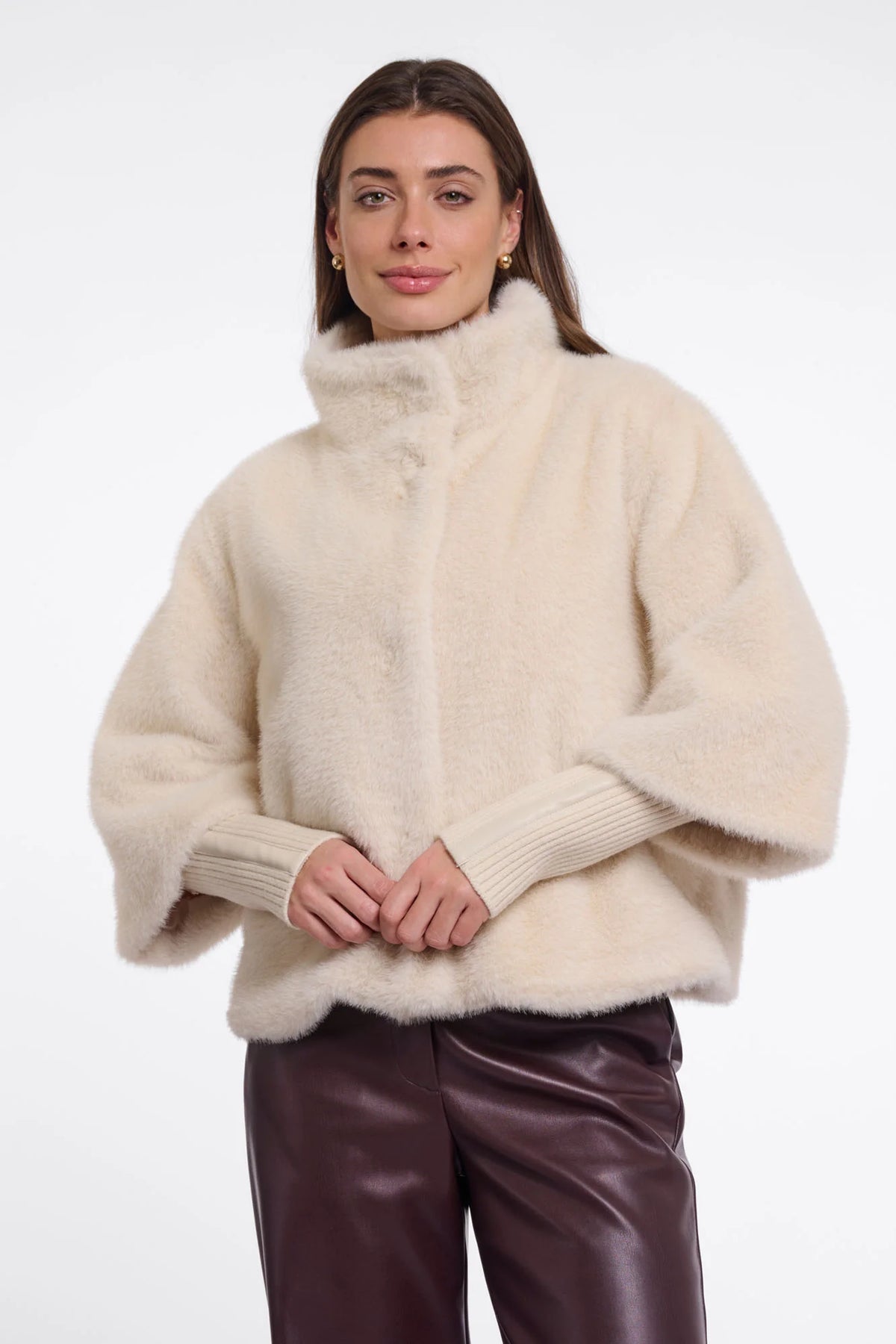 A woman wearing a cream-colored, fluffy faux-fur cropped jacket with a high mock neck and ribbed knit sleeves, paired with dark brown leather pants, against a plain white studio background.