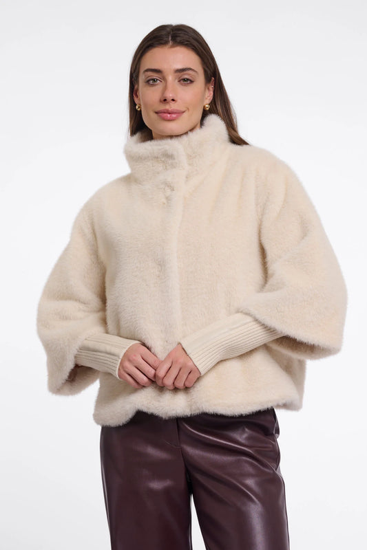 A woman wearing a cream-colored, fluffy faux-fur cropped jacket with a high mock neck and ribbed knit sleeves, paired with dark brown leather pants, against a plain white studio background.